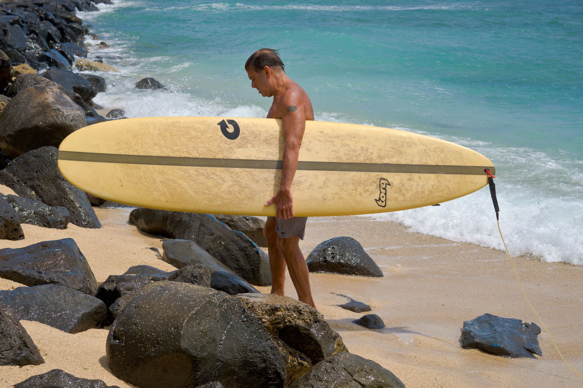 A senior man holding a longboard walks on a rocky beach with clear blue water.