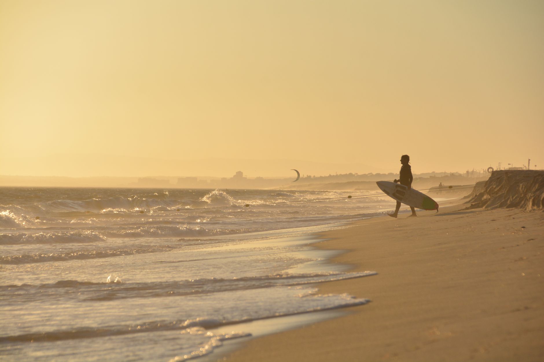 A surfer with a board walks along a serene beach at sunset, waves gently lapping the sand.